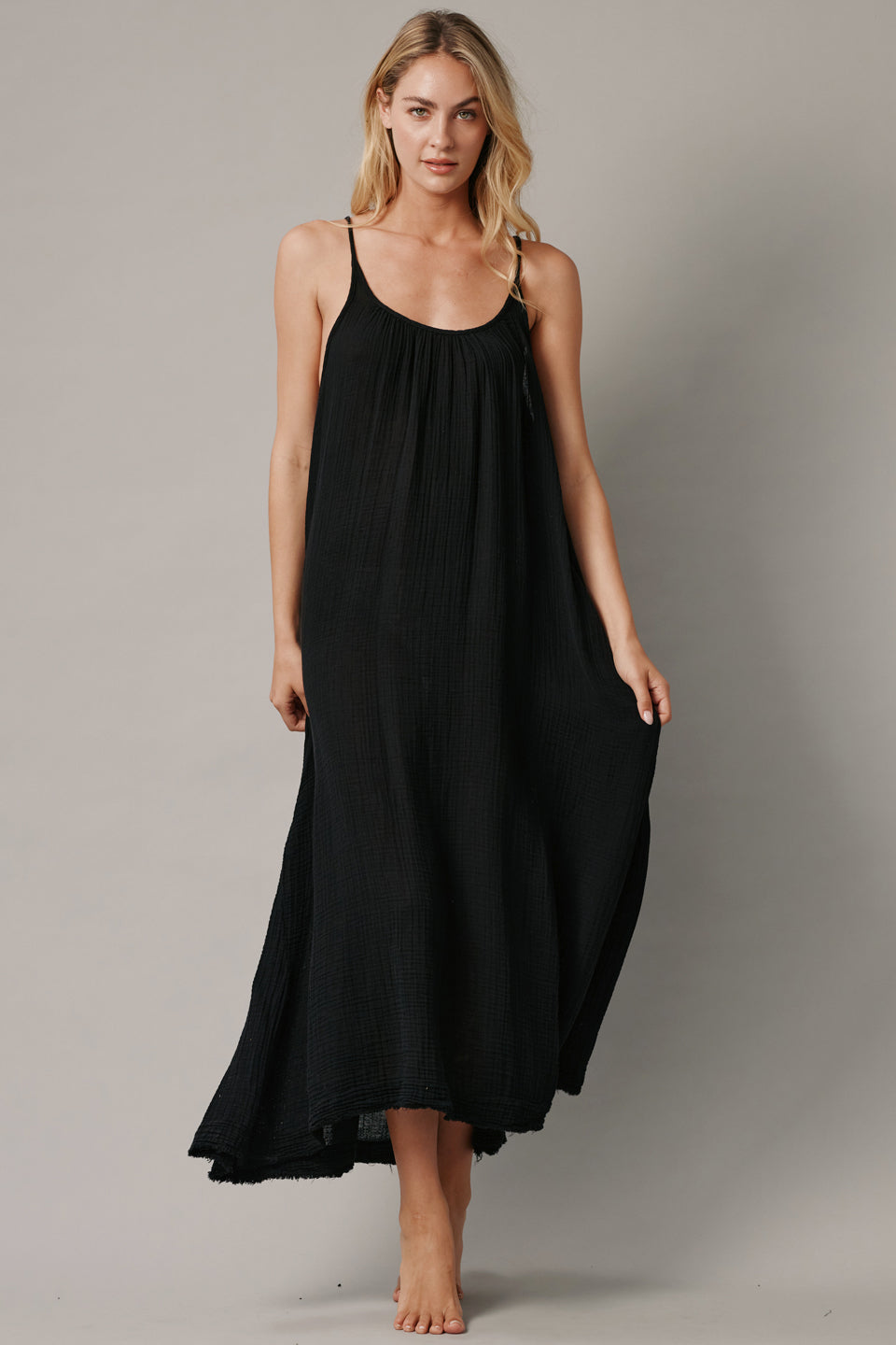 The full body front photo of a beautiful blonde woman wearing a TULUM black maxi dress in a lightweight gauze. She is standing on a grey canvas.