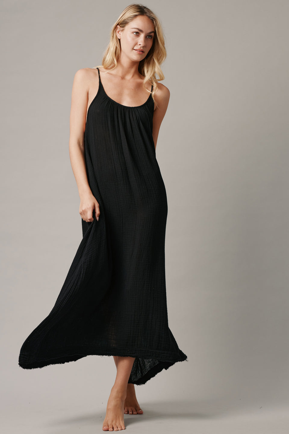 The full body with movement front photo of a beautiful blonde woman wearing a TULUM black maxi dress in a lightweight gauze. She is standing on a grey canvas.