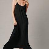 The full body with movement front photo of a beautiful blonde woman wearing a TULUM black maxi dress in a lightweight gauze. She is standing on a grey canvas.
