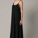 The full body side photo of a beautiful blonde woman wearing a TULUM black maxi dress in a lightweight gauze. She is standing on a grey canvas.