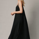 The full body with movement side photo of a beautiful blonde woman wearing a TULUM black maxi dress in a lightweight gauze. She is standing on a grey canvas.