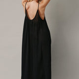 The full body back photo of a beautiful blonde woman wearing a TULUM black maxi dress in a lightweight gauze. She is standing on a grey canvas.