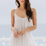 A cropped-in front photo of a beautiful brunette woman wearing a white maxi dress in a lightweight gauze, named TULUM, standing on a beach house balcony with the ocean in the background.