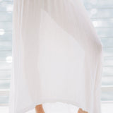 The lower half photo of a beautiful woman wearing a white maxi dress in a lightweight gauze, named TULUM, standing on a beach house balcony with the ocean in the background.