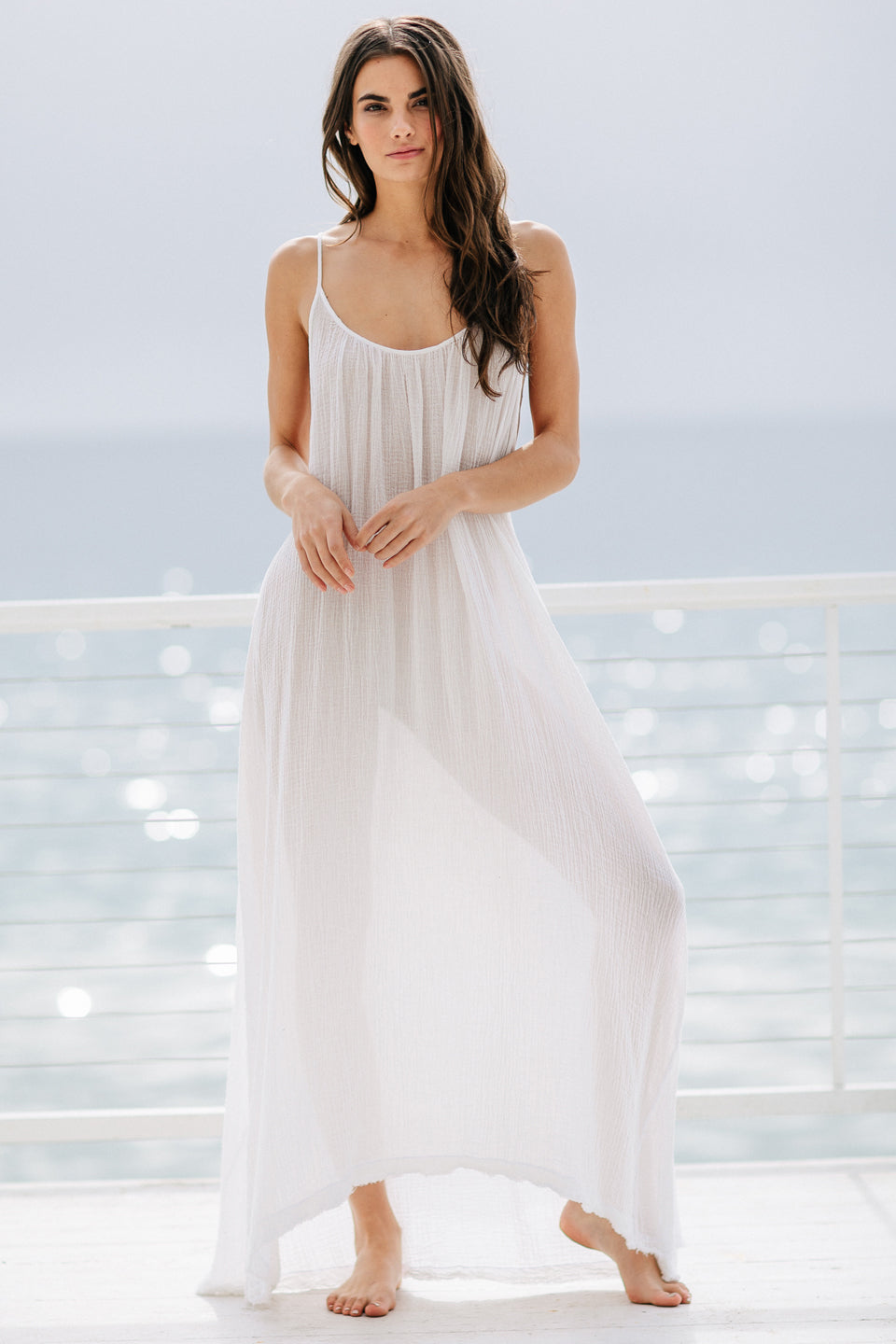 The full body front photo of a beautiful brunette woman wearing a white maxi dress called TULUM in a lightweight gauze, standing on a beach house balcony with the ocean in the background.