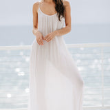 The full body front photo of a beautiful brunette woman wearing a white maxi dress called TULUM in a lightweight gauze, standing on a beach house balcony with the ocean in the background.