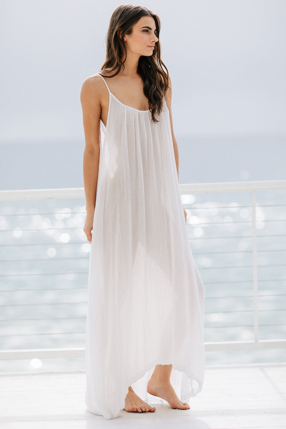 The full body 45 degree angle photo of a beautiful brunette woman wearing a white maxi dress in a lightweight gauze, named TULUM, standing on a beach house balcony with the ocean in the background.