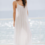 The full body 45 degree angle photo of a beautiful brunette woman wearing a white maxi dress in a lightweight gauze, named TULUM, standing on a beach house balcony with the ocean in the background.