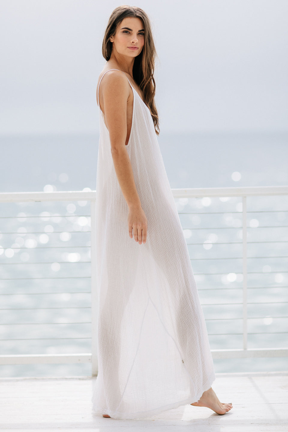 The full body side angle photo of a beautiful brunette woman wearing a white maxi dress in a lightweight gauze, named TULUM, standing on a beach house balcony with the ocean in the background.
