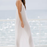 The full body side angle photo of a beautiful brunette woman wearing a white maxi dress in a lightweight gauze, named TULUM, standing on a beach house balcony with the ocean in the background.