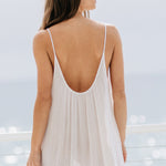 A cropped in back photo of a beautiful brunette woman wearing a white maxi dress in a lightweight gauze, named TULUM, standing on a beach house balcony with the ocean in the background.