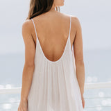 A cropped in back photo of a beautiful brunette woman wearing a white maxi dress in a lightweight gauze, named TULUM, standing on a beach house balcony with the ocean in the background.
