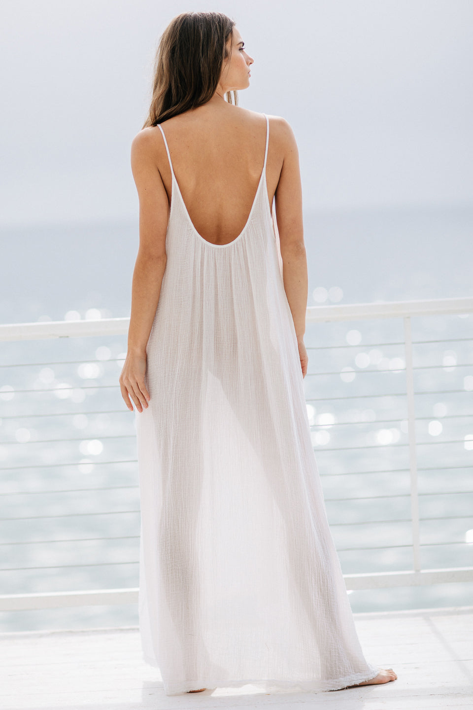 The full body back photo of a beautiful brunette woman wearing a white maxi dress in a lightweight gauze, named TULUM, standing on a beach house balcony with the ocean in the background.