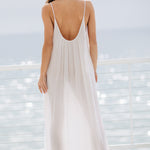 The full body back photo of a beautiful brunette woman wearing a white maxi dress in a lightweight gauze, named TULUM, standing on a beach house balcony with the ocean in the background.