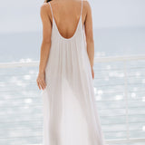 The full body back photo of a beautiful brunette woman wearing a white maxi dress in a lightweight gauze, named TULUM, standing on a beach house balcony with the ocean in the background.