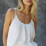 A blonde woman wearing a cotton white gauze tank top infornt of a grey sheet background. The tank top is slightly tucked in in the front.