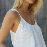 A cropped in 45 degree angle of the right side of a blonde woman wearing a cotton white gauze tank top in front of a grey sheet background. The tank top is slightly tucked in in the front.
