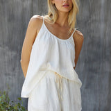 Cropped slightly above the knee, a photo of a blonde woman wearing a cotton white gauze tank top in front of a grey sheet background. The tank top is slightly tucked in in the front with white shorts.