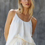 Cropped in front photo of a blonde woman wearing a cotton white gauze tank top in front of a grey sheet background. The tank top is slightly tucked in in the front with white shorts.