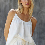 Cropped in front photo of a blonde woman wearing a cotton white gauze tank top in front of a grey sheet background. The tank top is slightly tucked in in the front with white shorts.