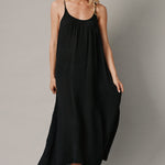 The full body front photo of a beautiful blonde woman wearing a TULUM black maxi dress in a lightweight gauze. She is standing on a grey canvas.