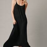 The full body with movement front photo of a beautiful blonde woman wearing a TULUM black maxi dress in a lightweight gauze. She is standing on a grey canvas.