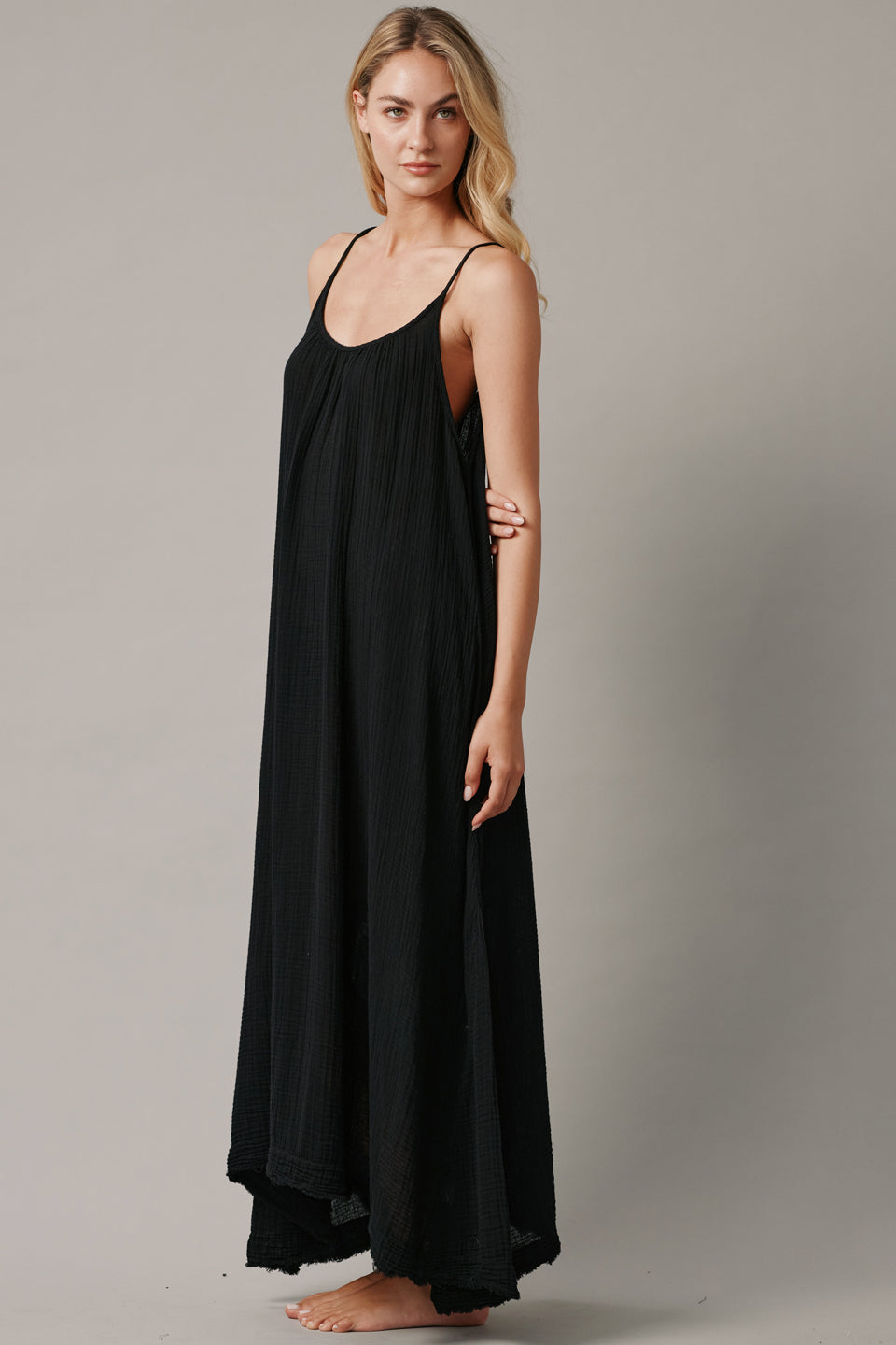 The full body side photo of a beautiful blonde woman wearing a TULUM black maxi dress in a lightweight gauze. She is standing on a grey canvas.