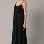 The full body side photo of a beautiful blonde woman wearing a TULUM black maxi dress in a lightweight gauze. She is standing on a grey canvas.