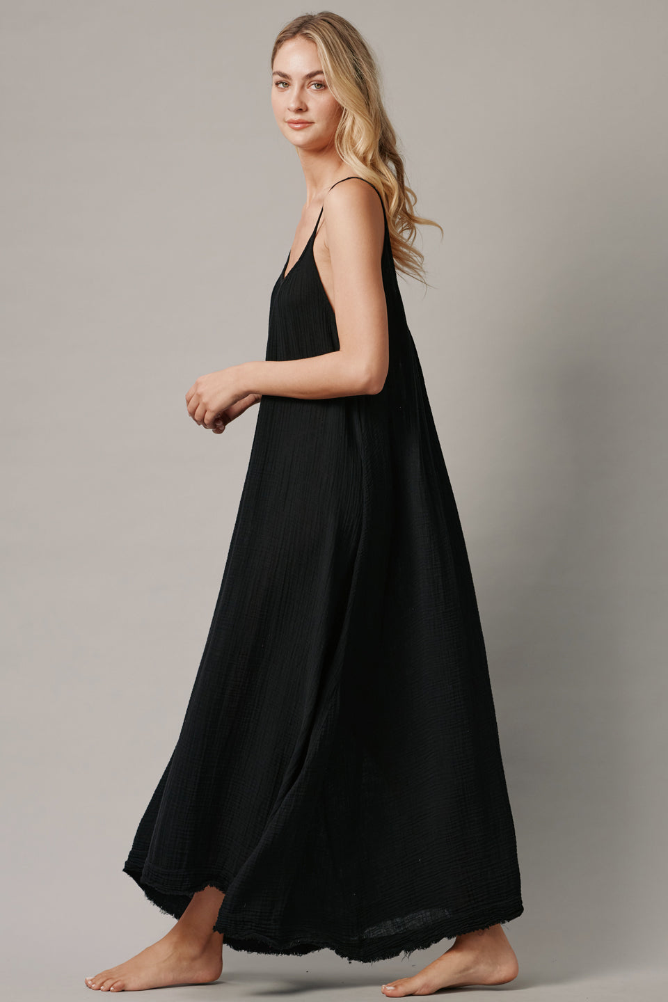 The full body with movement side photo of a beautiful blonde woman wearing a TULUM black maxi dress in a lightweight gauze. She is standing on a grey canvas.