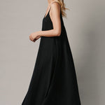 The full body with movement side photo of a beautiful blonde woman wearing a TULUM black maxi dress in a lightweight gauze. She is standing on a grey canvas.