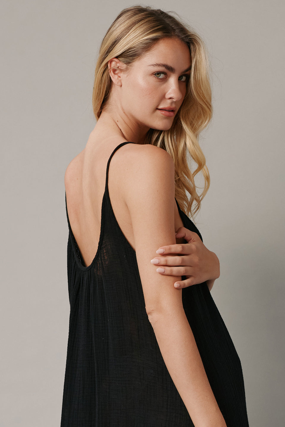 The close up back angled photo of a beautiful blonde woman wearing a TULUM black maxi dress in a lightweight gauze. She is standing on a grey canvas.