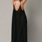 The full body back photo of a beautiful blonde woman wearing a TULUM black maxi dress in a lightweight gauze. She is standing on a grey canvas.